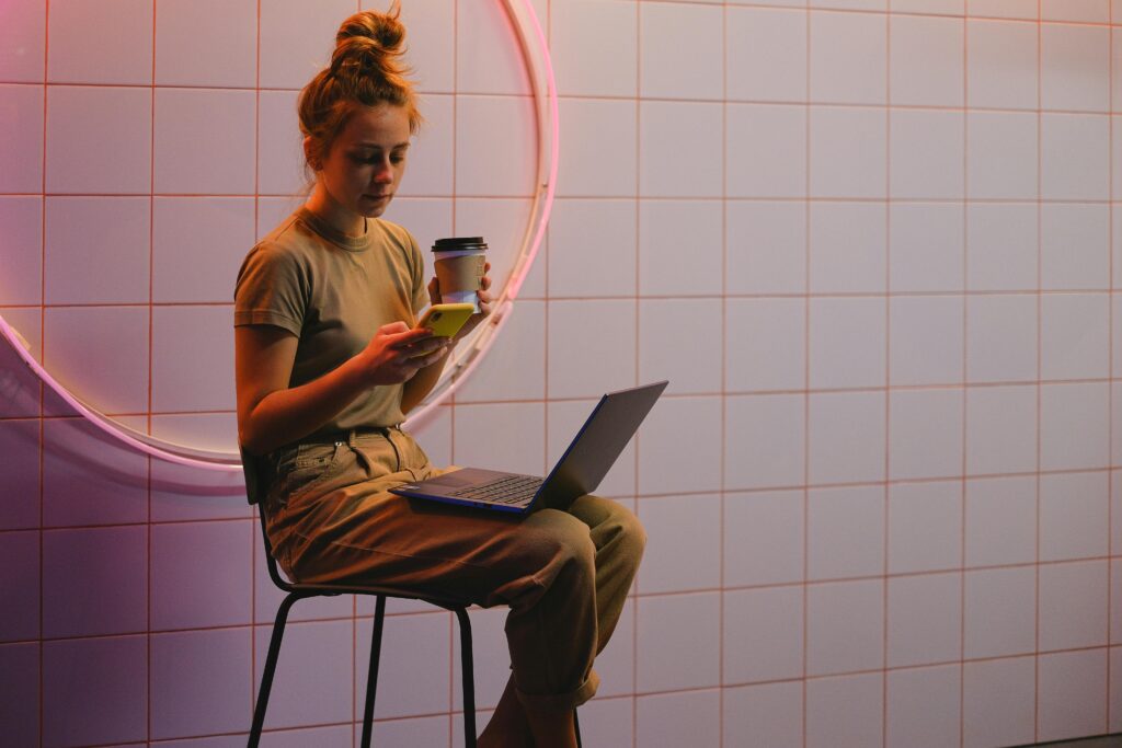 Young woman multitasks with phone and laptop in a trendy cafe with neon lighting.