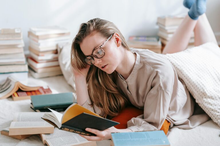A young woman relaxing at home, engrossed in a book amid piles of literature.
